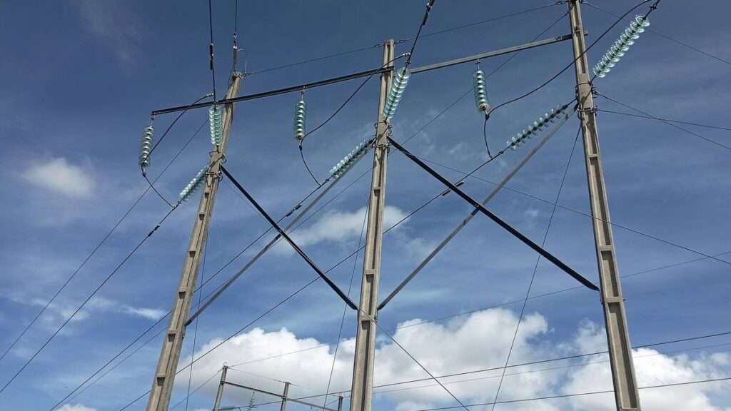 Dos torres de transmisión eléctrica con cadenas de aisladores de vidrio, contra un cielo azul con nubes.