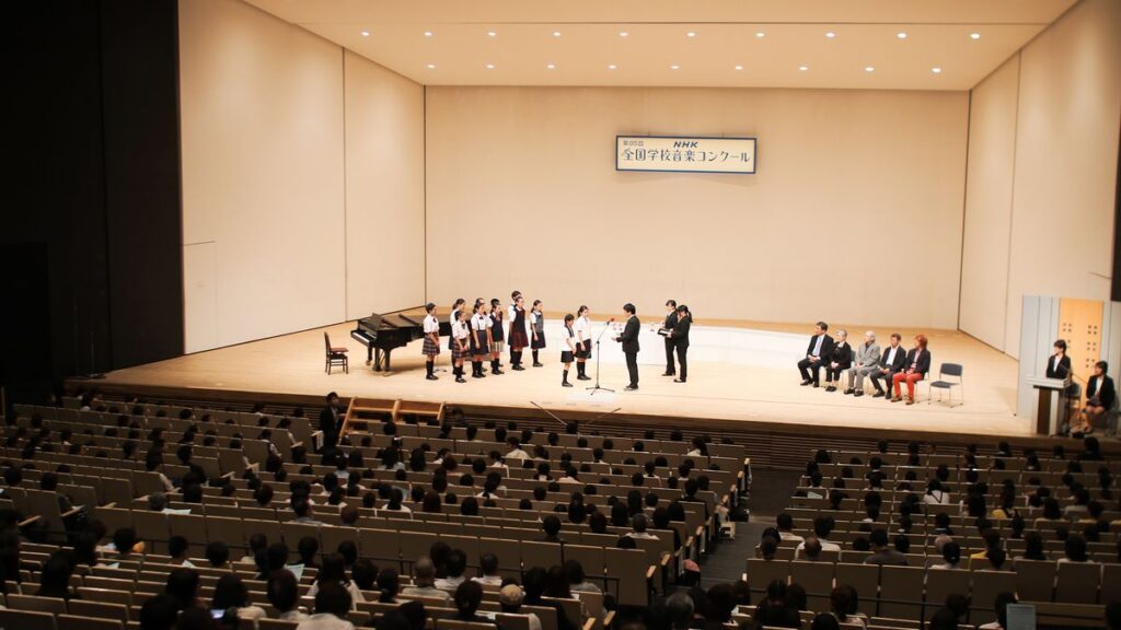Un coro escolar con uniforme de colegio canta sobre el escenario de un auditorio, frente a la platea con público.