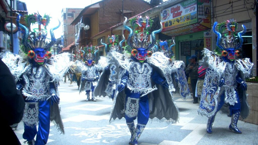 Bailarines de diablada con máscaras de diablo de colores y trajes azules con plateado desfilan por una calle.