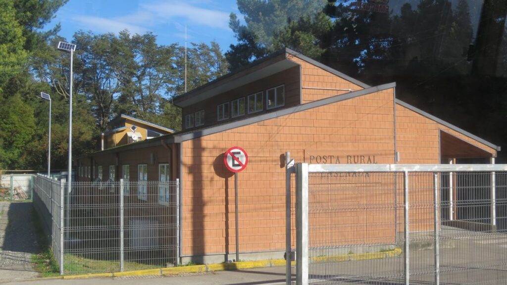 Edificio de una posta rural de un piso y medio, con muros de tejuela color naranjo y rejas metálicas al frente, rodeado de árboles.