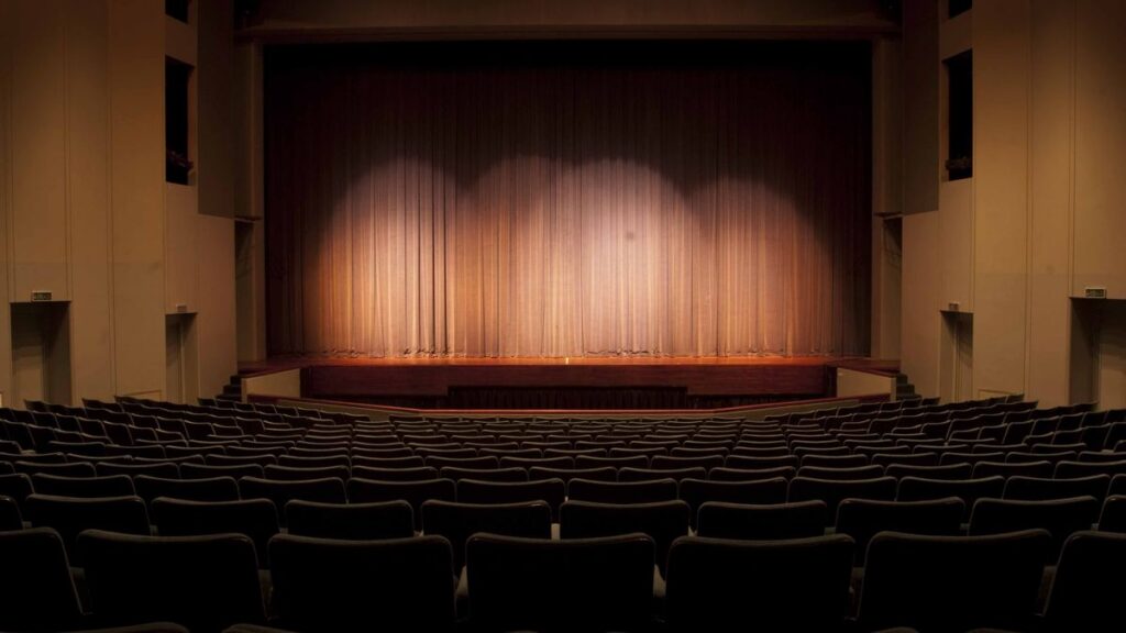 Escenario de un teatro con el telón cerrado iluminado por focos, frente a la platea vacía.
