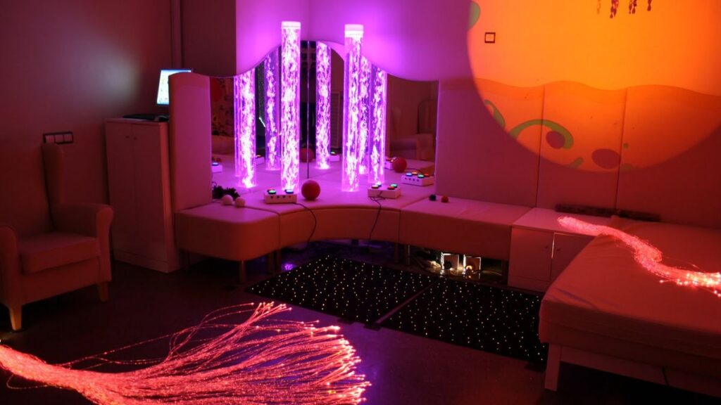 Sala sensorial en penumbra con columnas de burbujas iluminadas en violeta, cojines blancos, una alfombra con luces y haces de fibra óptica en el suelo.